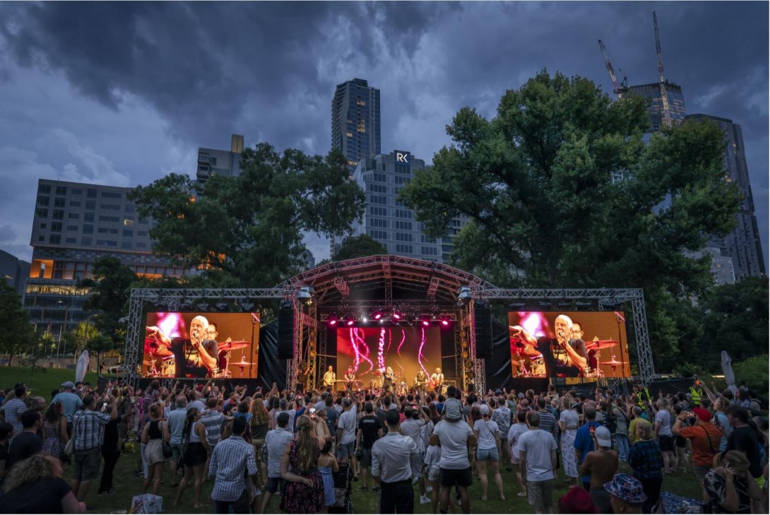 Lots of people gathered outdoors in a park with tall city buildings in the background, in front of a stage with big TV screens showing a concert.