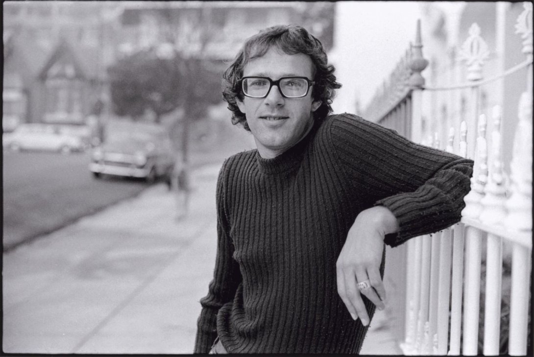 A black and white image of a man in glasses and a dark jumper, leaning against a fence. In the background is a street with a car parked on it.