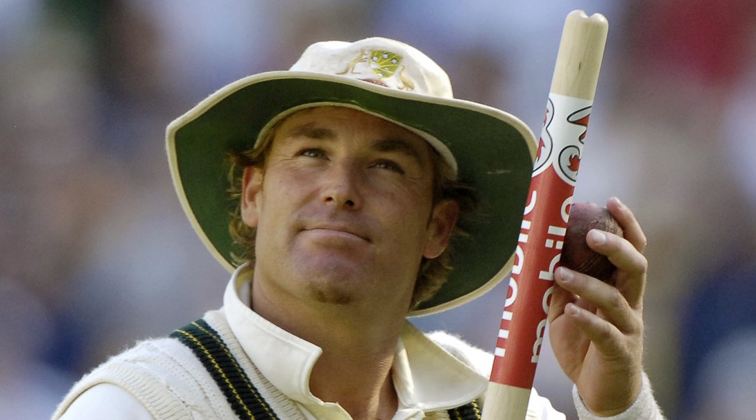 A man in cricket whites and a hat holding a cricket stump and looking upwards.