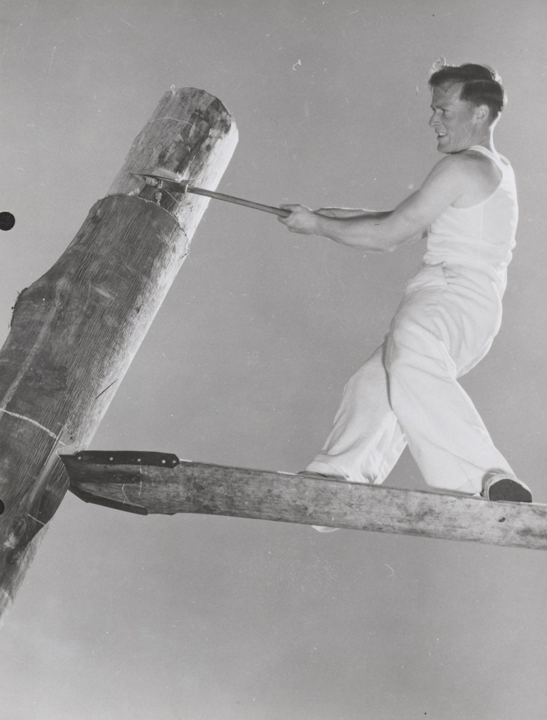 A man in a white singlet and pants standing on a flat piece of wood which is stuck into a tall circular piece of wood. 