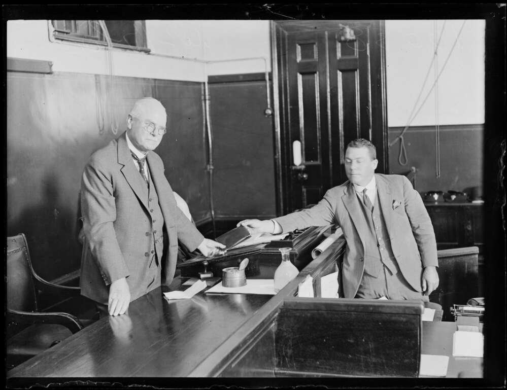 Older man in a three-piece suit standing behind very long desk being handed a book by a man on the other side of the desk who is also in a three piecesuit.