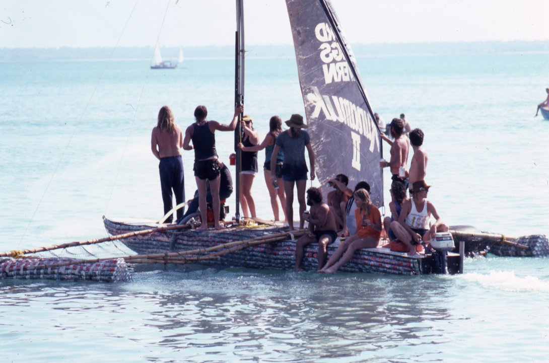 A boat made of drink cans in the water with approximately 10 people standing or sitting on the boat. 