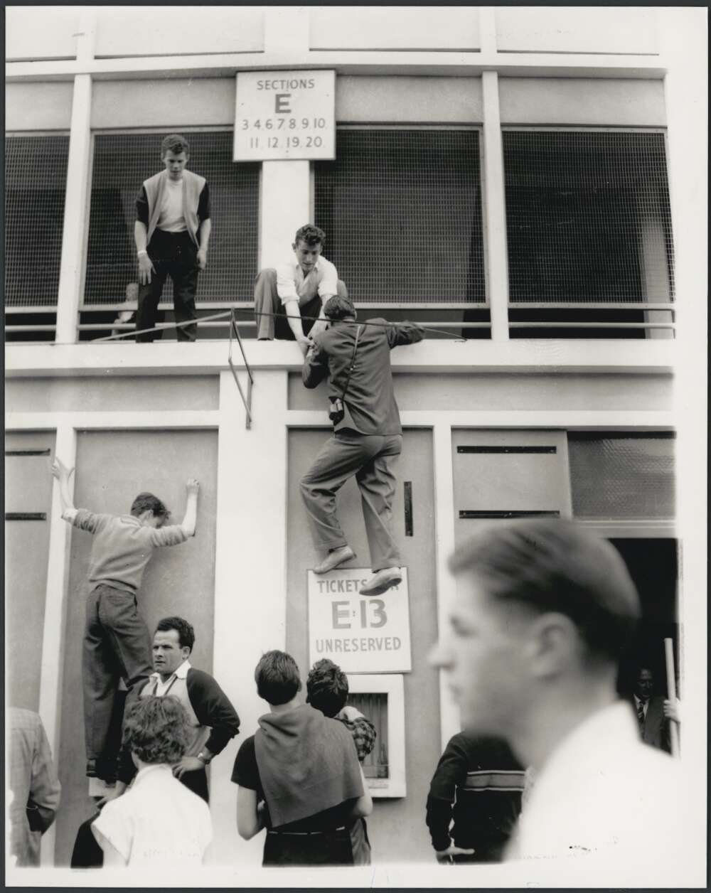 Young men helping eachother climb a wall with signs reading 'Sections E', while other people stand around or walk past
