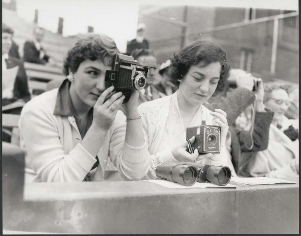 Two women leaning over railing in front of stands in sports arena using old cameras to capture the sports taking place