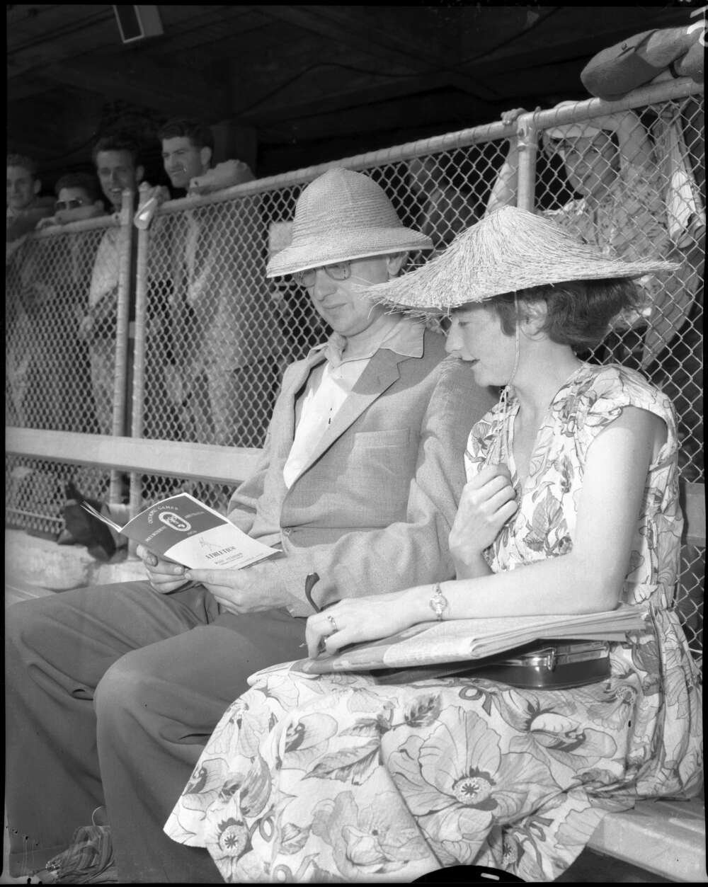 Man and woman sitting in the stands of a sports arena wearing nice clothes andsunhats