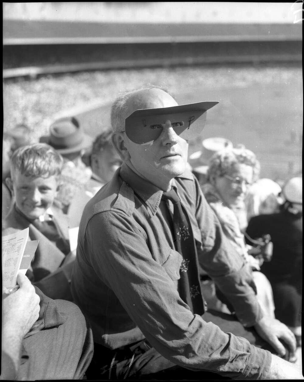 Man in the crowded stands of a sports arena wearing shirt and tie and cardboard visor that sits around his eyes and extends forward from his forehead, tied around the back of his head