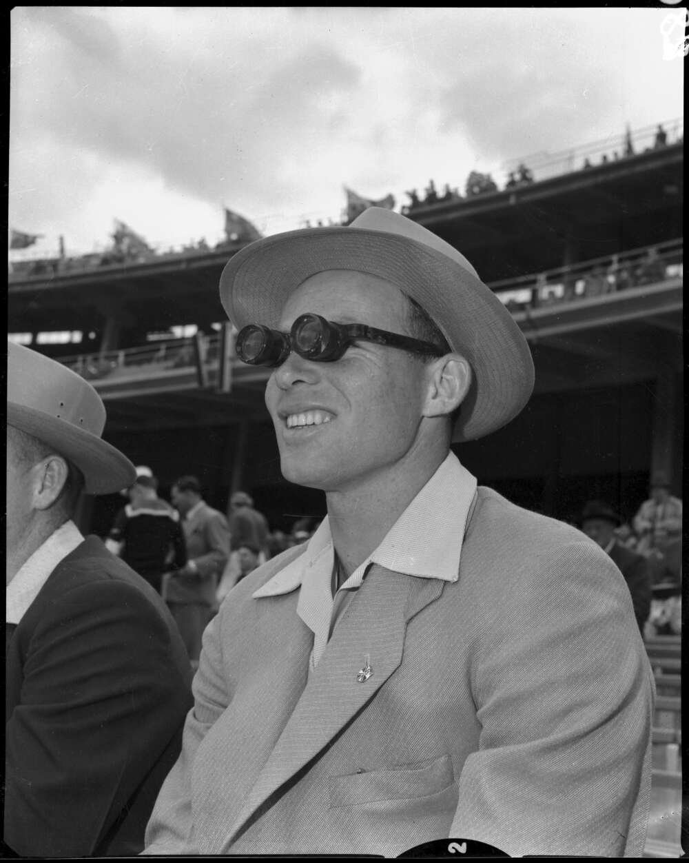 Man sitting in stands of sports stadium in suit and sunhat using binocular glasses