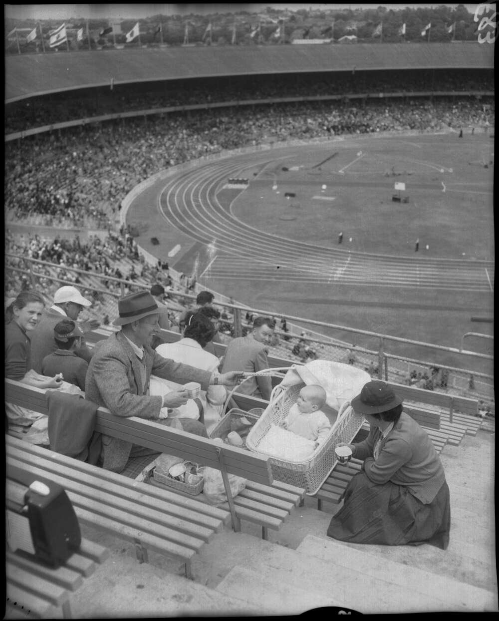 Family of three sitting in the crowded stands of a sports stadium while various events take place on the track and field. The parents seem to be enjoying a warm beverage while looking at their baby who lies in a stroller.