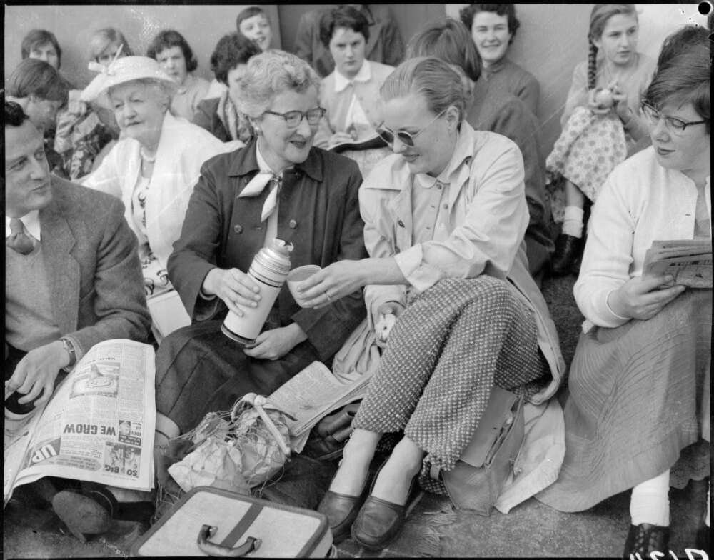 Two women sitting amongst other spectators sharing lunch and pouring a drink from a thermos
