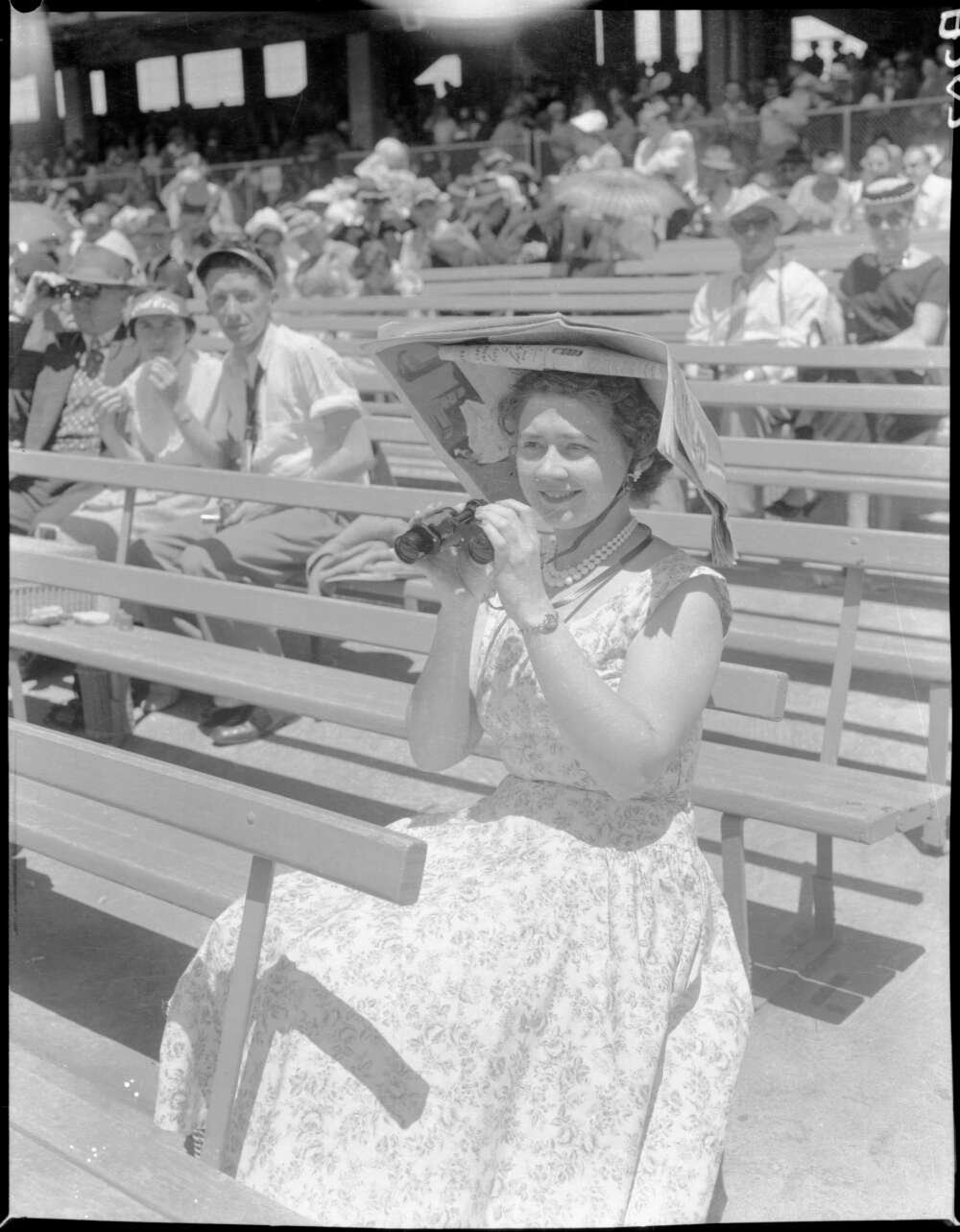 Woman sitting in the stands of a sports arena in a nice floral dress, holding up binoculars and wearing a large sunhat made out of newspapers