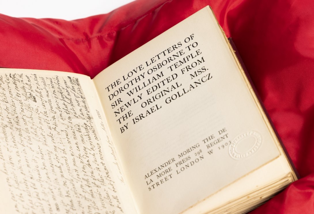 Photo of a rare book on a red cushion with an image of a manuscript on the left and a printed title page on the right