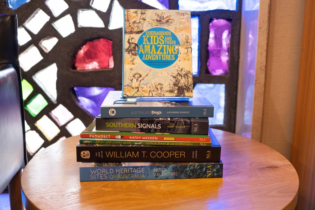 Stack of books on a round wooden table in front of colourful stained glass windows. The books are various sizes and colours