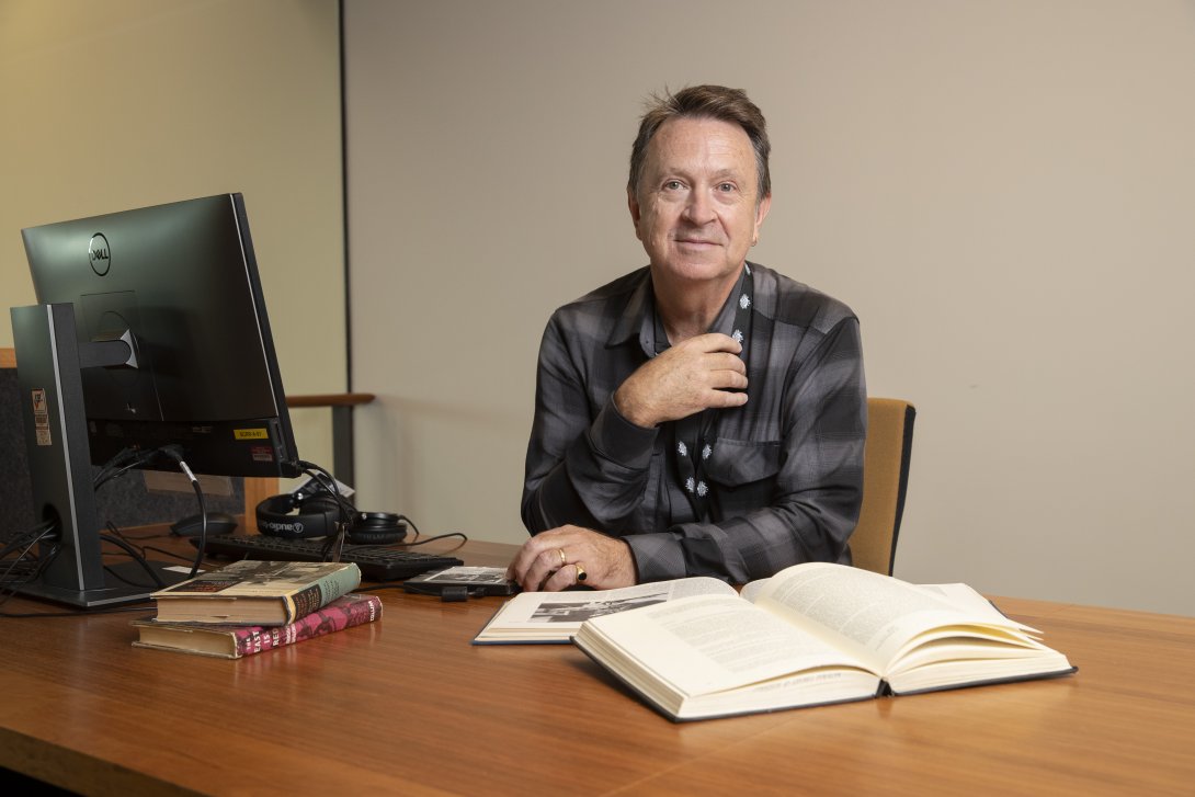 A man wearing a black and grey checkered shirt sitting at a desk with a computer and several books resting on it. He is leaning on the desk, facing forward and giving the camera a small smile