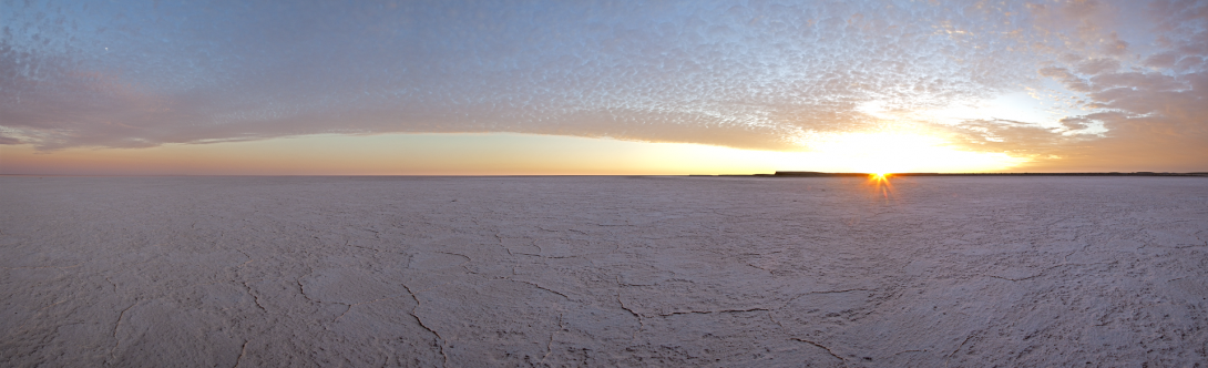 Wide photo of a dry salt lake that stretches as far as the eye can see. The sky is varying shades of orange, blue and purple as the sun is sitting just on the horizon