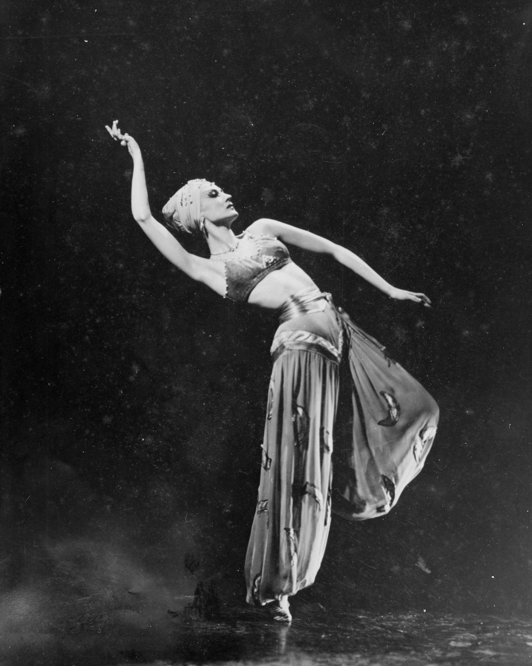 Woman in front of a black background, wearing a headdress and dance costume, posed so she's standing on one leg, leaning to the side, and lifting one arm above her head
