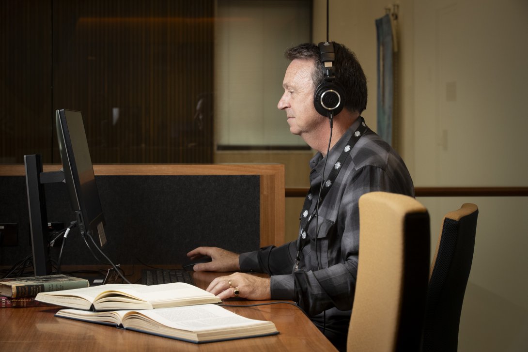 Man in a grey and black checkered shirt and headphones sitting at a desk, using a computer. To his left are two open books.