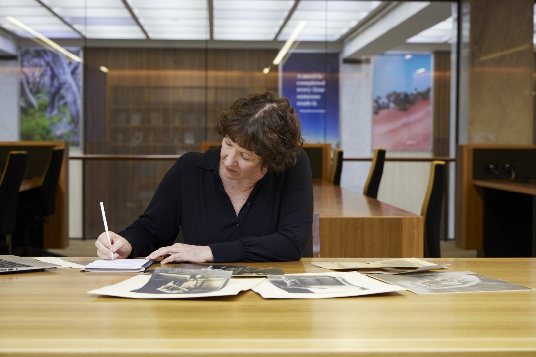 Woman with short, curly brown hair wearing a black top sitting at a table covered in old photos and manuscripts, writing in a notebook