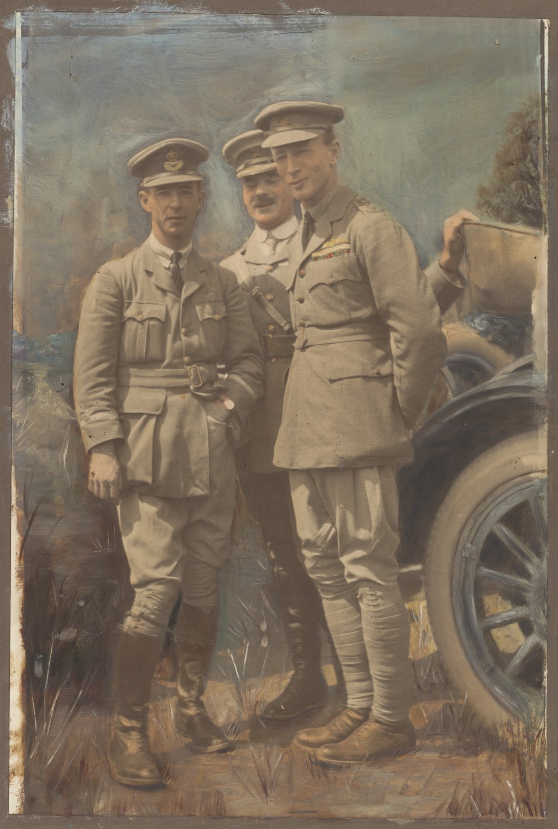 Three men in military uniform, including hats, standing closely together in front of a car. 