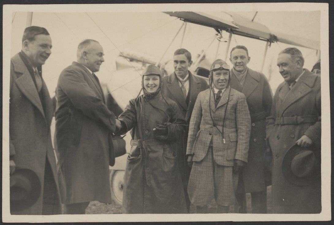Female pilot in long coat smiling and shaking hands with a large man. Around them are 5 other smiling men. In the background, a wing of a plane can be seen
