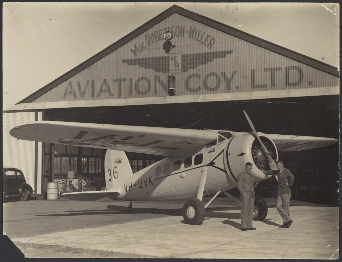 Two men standing and leaning against a small passenger plane in front of an aircraft hangar