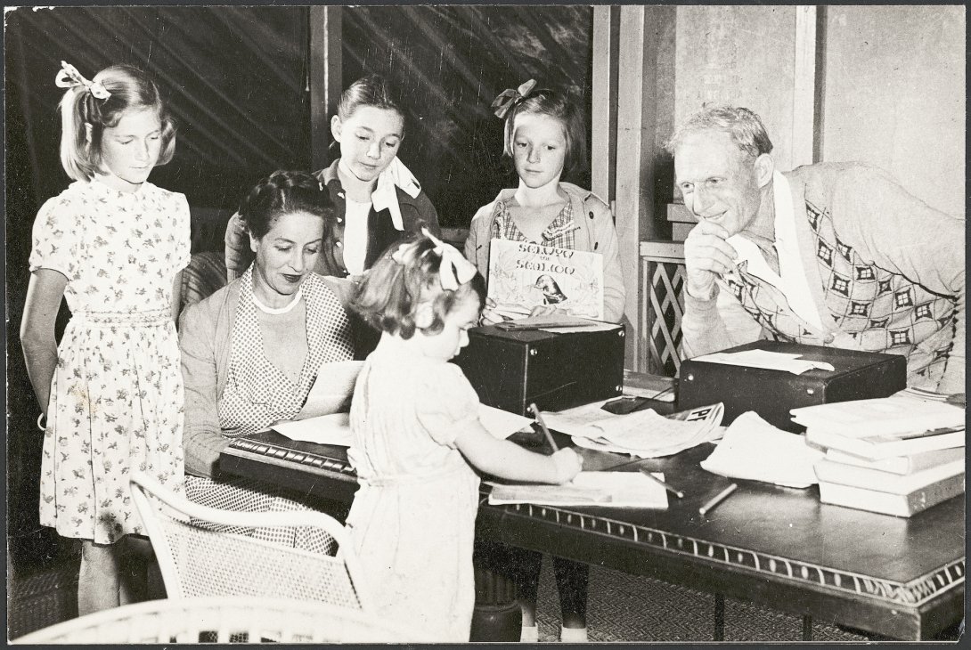 Family with mother, father and four young daughters sitting around a table with papers and boxes on it. The daughters al have bows in their hair and they all look content