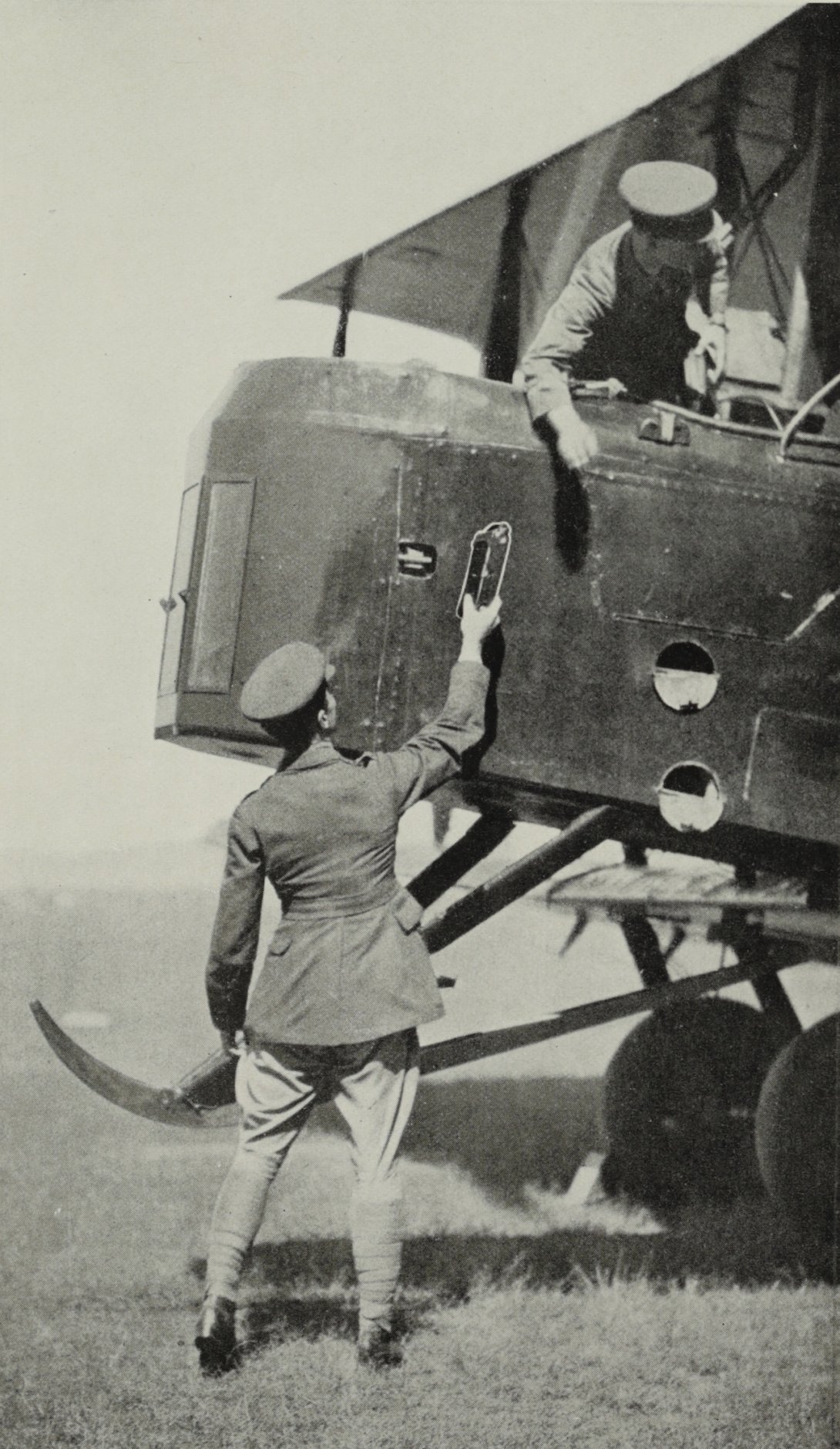 Man in military uniform standing next to front of plane, reaching up with camera in his hand while man in cockpit reaches down to grab it