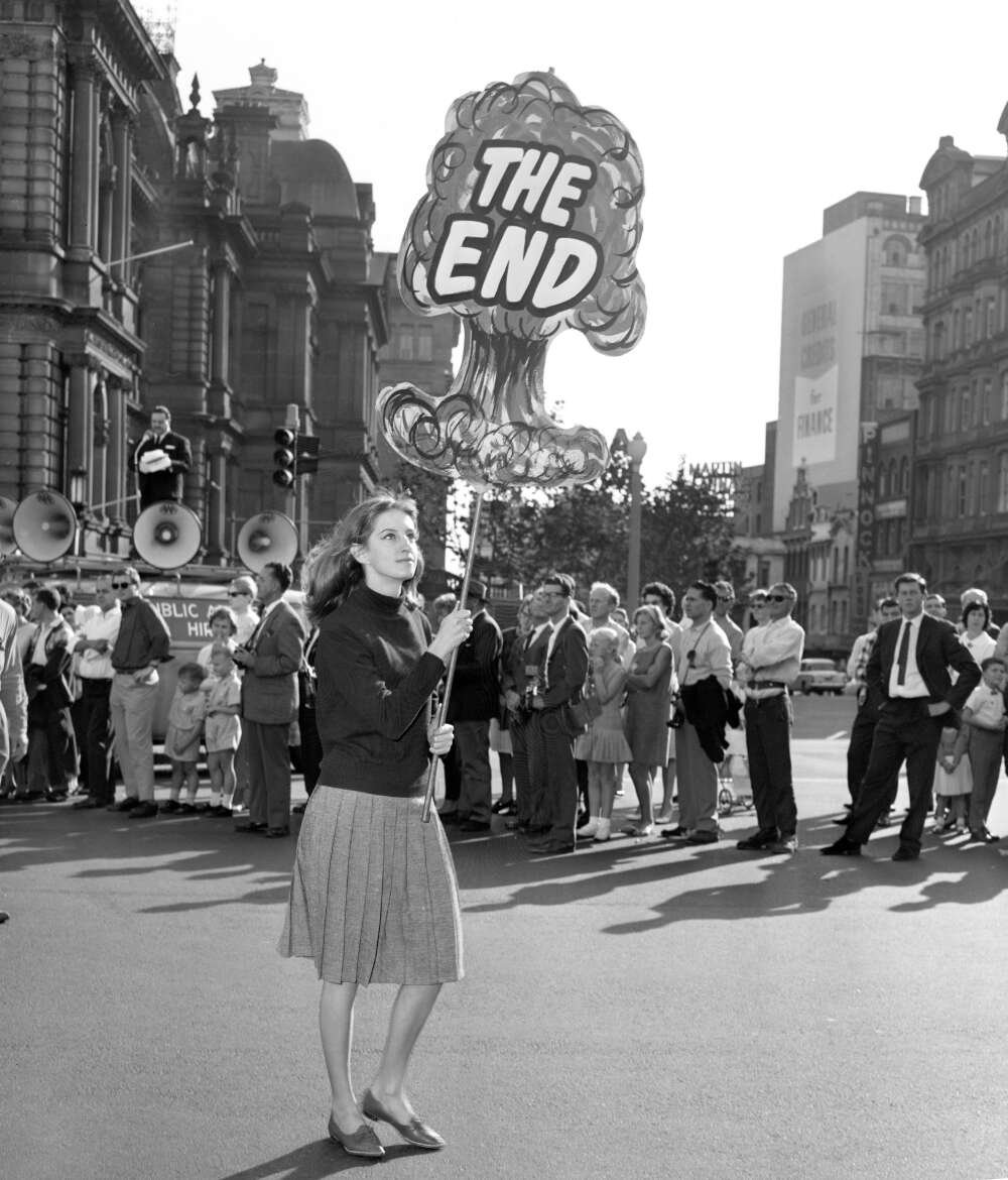 Young woman walking through a parade with a sign shaped like a mushroom cloud with text saying 'THE END'
