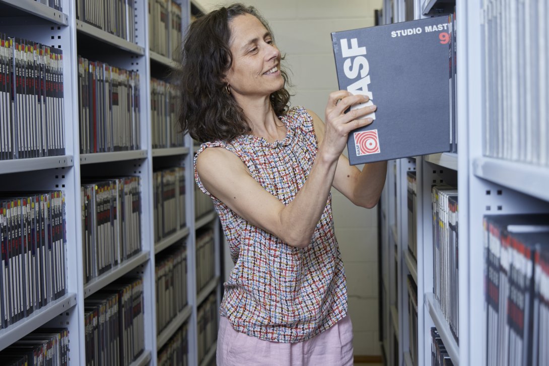 Woman with curly black hair standing by a shelf of black folders, putting one back on the shelf