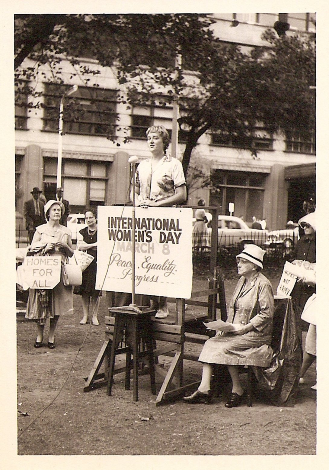 Woman on small wooden platform speaking into a microphone. In front of her is a sign that reads 'International Womens Day. March 8. Peace. Equality. Progress.' A few women stand and sit around her listening.