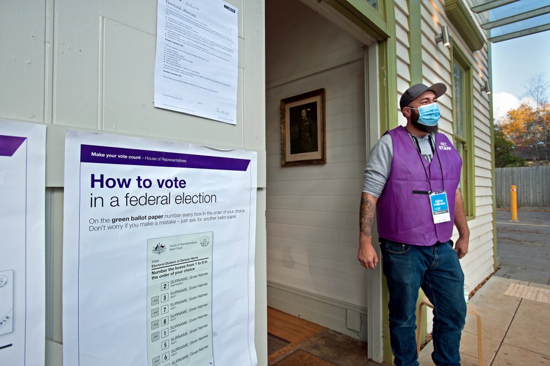 A man wearing a purple “Australian Electoral College Vest” stands beside the door of a polling place for the 2022 Federal Election