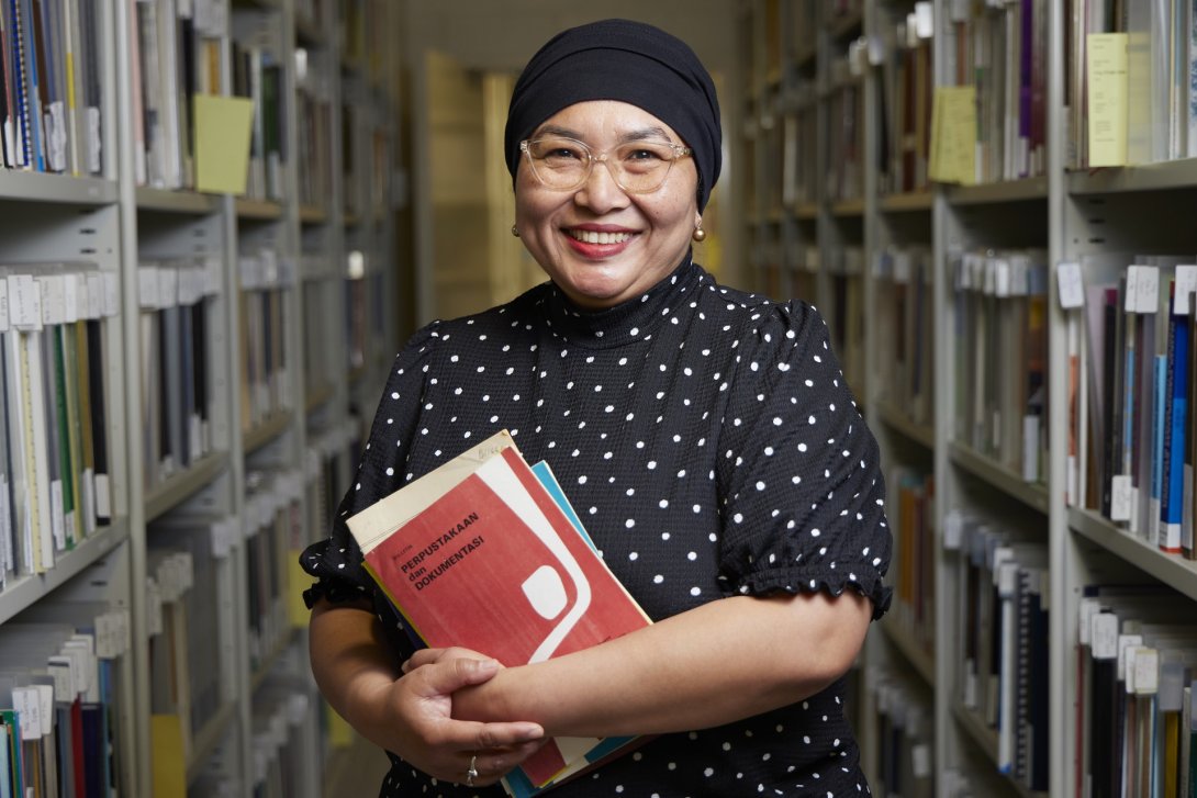 Woman wearing a dot print shirt, glasses and a black head scarf holding some booklets in an aisle of shelves