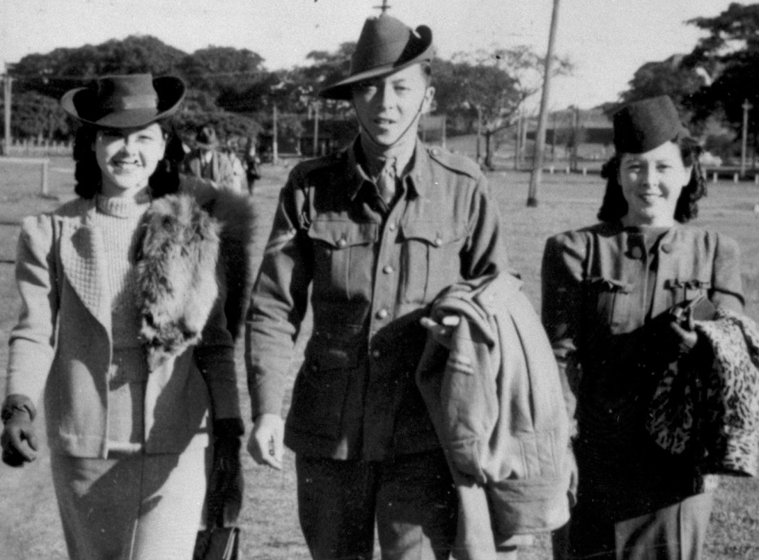 Two well-dressed young women and one young man in military uniform smiling and walking through a park