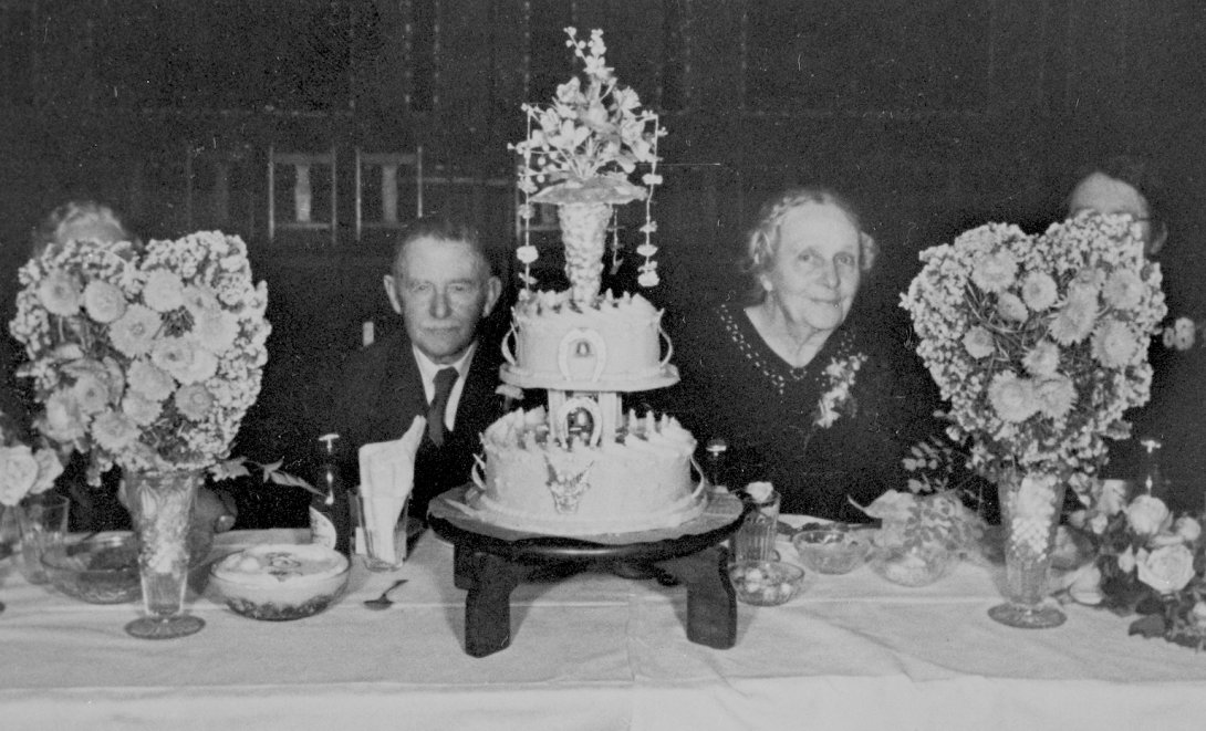 Husband and wife sitting at a table with a large cake and vases of flowers in front of them