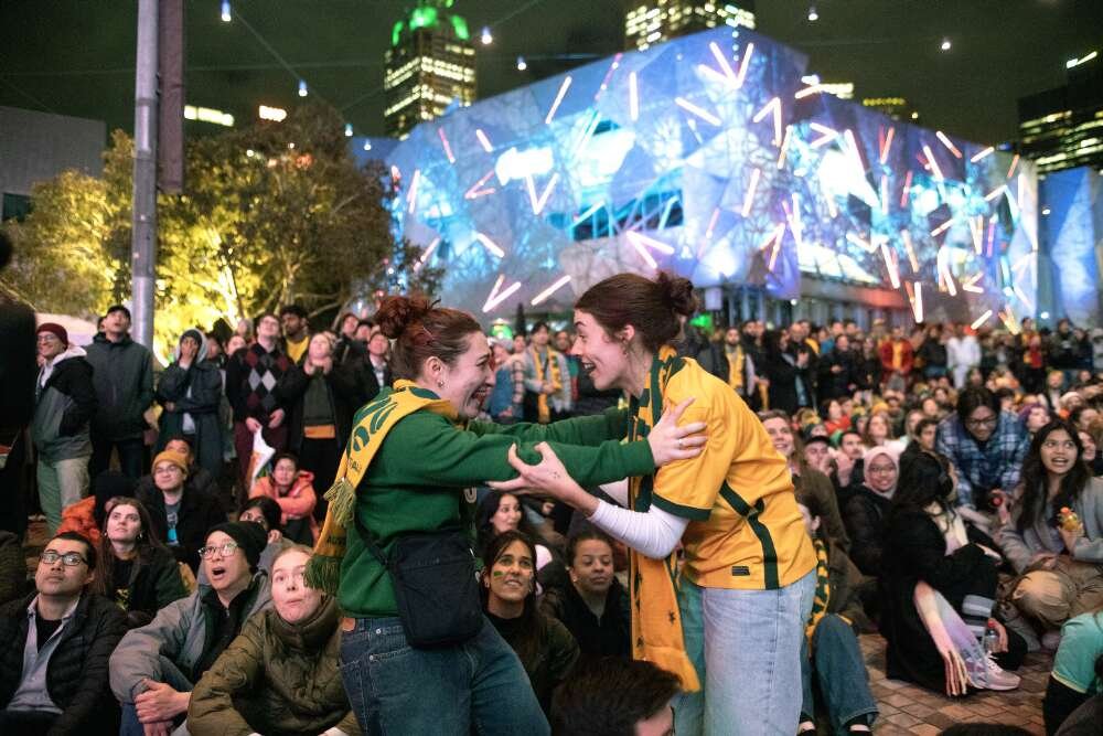 Two woman in green and gold clothing embracing excitedly in front of a crowd.