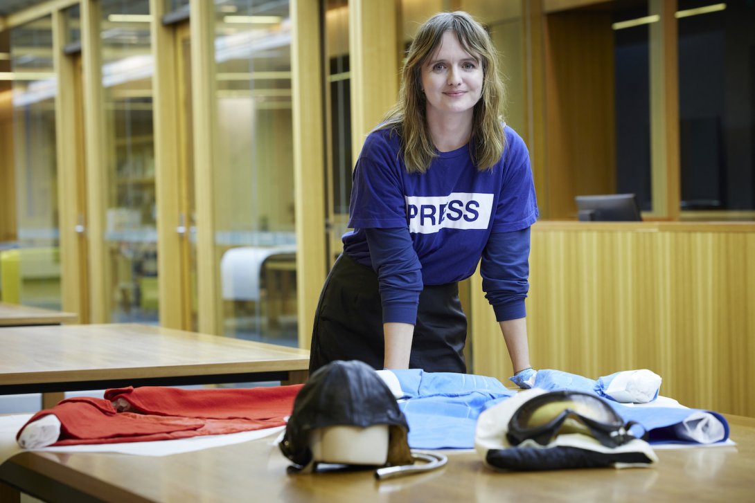 Dr Jo Langdon standing and leaning on a table in the Special Collections Reading Room with a Royal Perth Hospital Nurses uniform worn including a red cape and two pilot's helmets and goggles on it