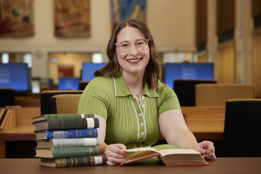 Zoe Smith, an NLA Scholar, sitting at a table in the Main Reading Room with a pile of books and smiling