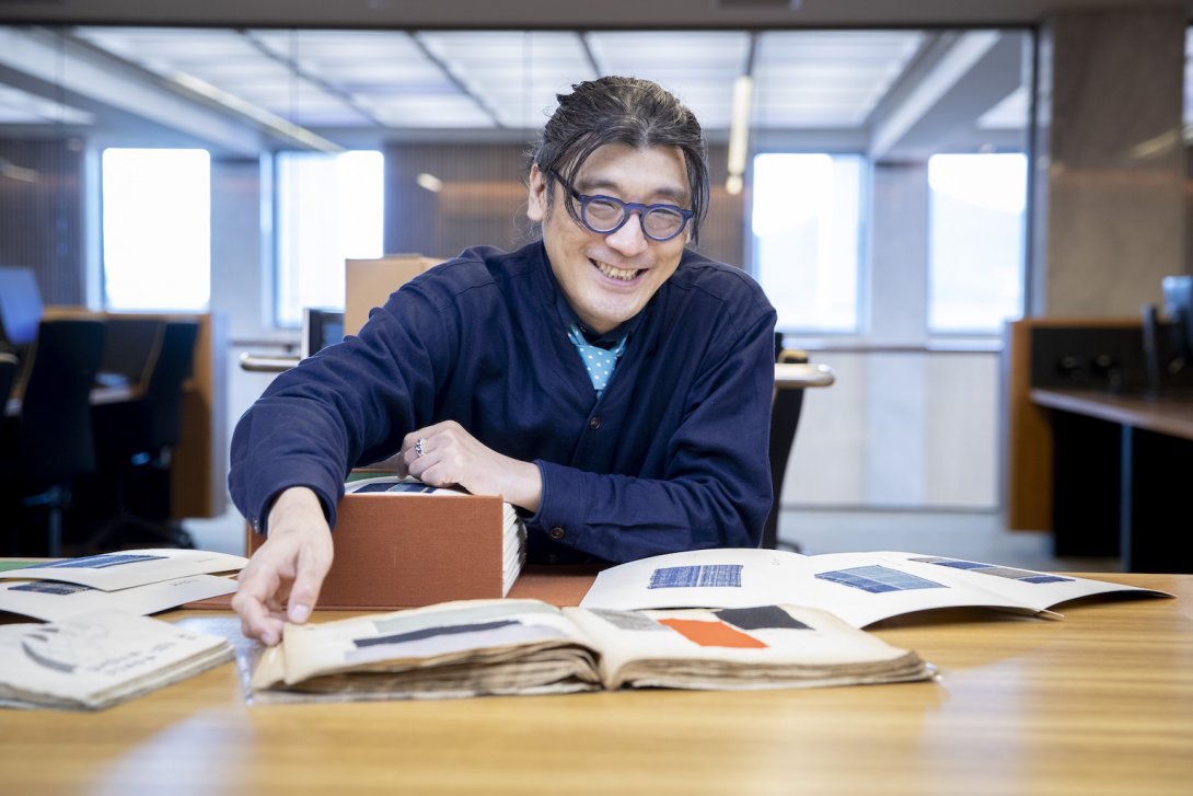 Dr Tets Kimura smiling and sitting at a table in the Special Collections Reading Room with material relating to Japanese fashion spread out in front of him