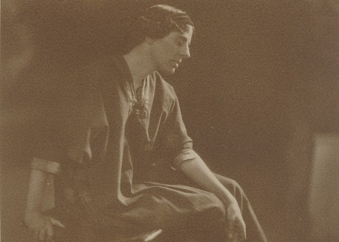 Sepia photograph of Marion Mahony Griffin, seated and in profile, with her back arm resting on her knees and front arm on the stool she is sitting on. She wears a long, loose dress.